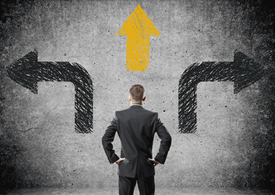 Businessman standing in front of concrete wall with three arrows stenciled on it, one left, one right and one up.