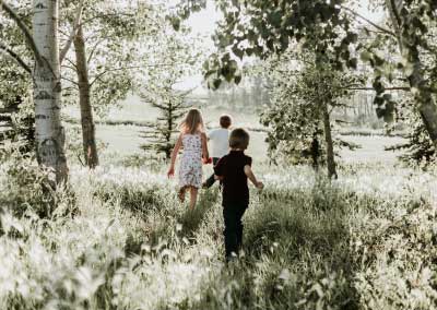 A girl dressed in white and two boys, one dressed in white and one in black walking out of a forest into a clearing