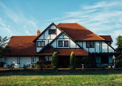 Tudor styled ranch house with orange-brown roof, white facades, and two garages. It has a bushy tree on each side of the door.