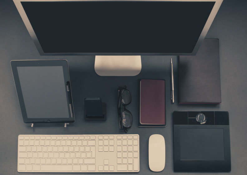 An array of business devices (tablet, phone, glasses, planner, notebook, keyboard, mouse, trackpad, and computer screen) arranged on a desk